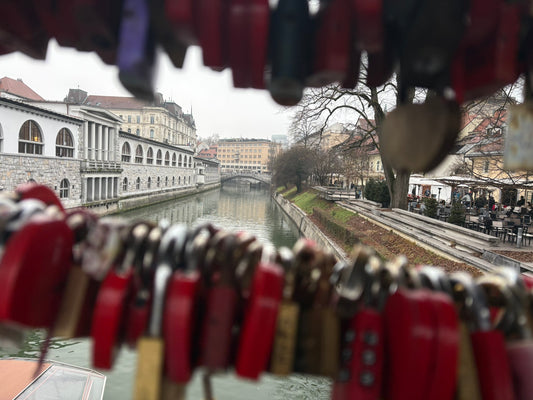 Eine Aufnahme aus der Perspektive eines Betrachters auf einer Brücke, eingerahmt von zahlreichen Liebesschlössern im Vordergrund.
Vordergrund: Eine Vielzahl von bunten, überwiegend roten und messingfarbenen Vorhängeschlössern hängt dicht an dicht an Drahtseilen. Die Schlösser sind leicht unscharf (Bokeh-Effekt), wodurch der Fokus auf den Hintergrund gelenkt wird.
Mittelgrund: Der ruhige Fluss Ljubljanica fließt zentral durch das Bild. Auf der linken Seite erstreckt sich das markante, weiße Arkadengebäude