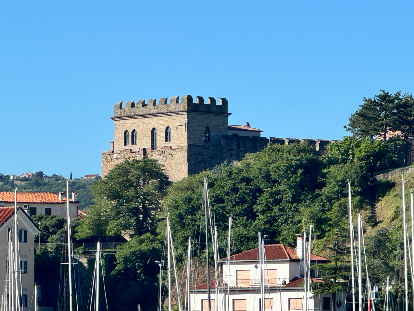 Blick vom Hafen in Muggia auf die Burg von Muggia. Im Vordergrund sieht man die Masten von Segelbooten, dahinter viele Bäume mit grünen Blättern und dahinter die Burg von Muggia mit vier Fenstern zum Meer und auf dem Dach 7 Zinnen.