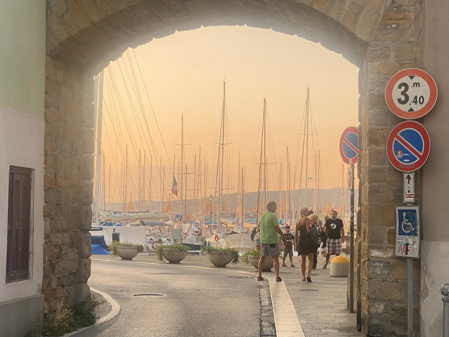 Blick durch ein altes Stadttor auf den Hafen von Muggia in der orangefarbenen Abendsonne. Rechts im Vordergrund drei Verkehrszeichen (Max. Höhe 3,4 Meter, Halten verboten und ein Schild für Rollstuhlfahrer). Im Hintergrund auf der rechten Seite eine Gruppe von Menschen - 5 Erwachsene und ein Kind.