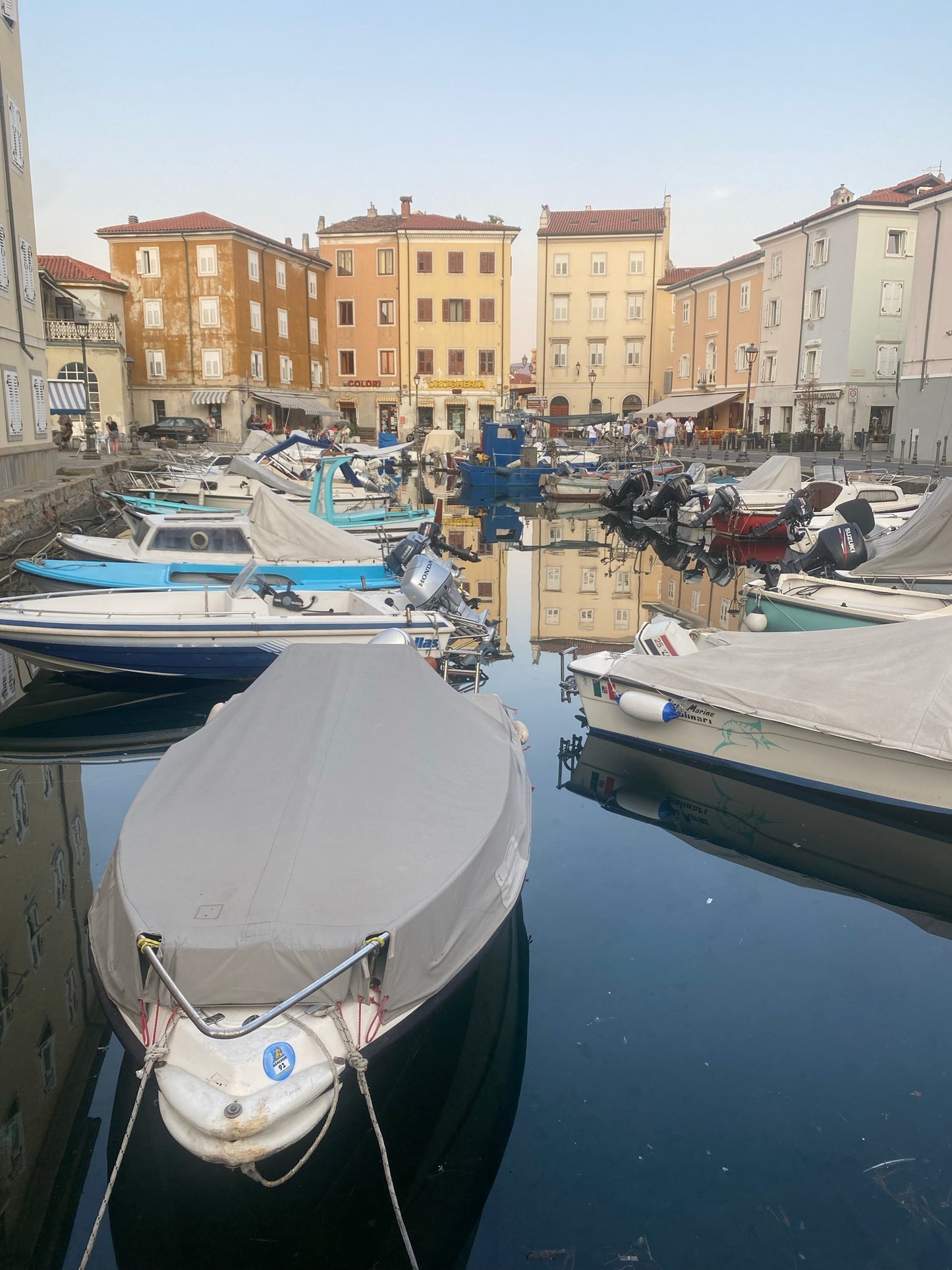 Blick auf den Hafen von Muggia (Mandracchio) mit bunten Schiffen auf dem blauen Meer in der Abendsonne und im Hintergrund bunte Häuser in orange, gelb und weiß, die sich zwischen den Booten im Meer spiegeln.