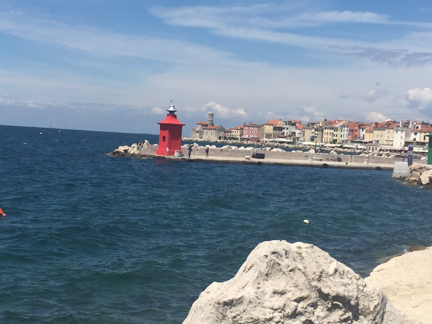 Aufnahme von Piran, im Vordergrund Steine und das Meer, dahinter ein kleiner roter Leuchtturm und dahinter die Altstadt von Piran