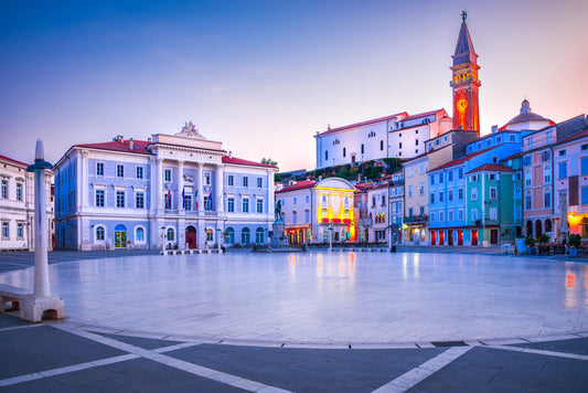 Tartini Platz in Abendstimmung mit Beleuchtung der Häuser und der St. Georgs-Kirche im Hintergrund. Im Vordergrund ist der runde, steinerne Teil des Platzes zu sehen.