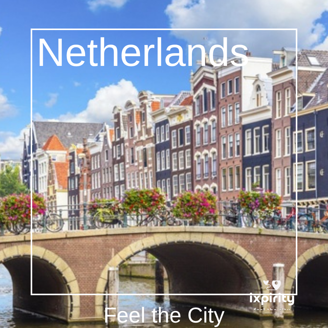 Eine Amsterdamer Gracht mit schmalen Giebelhäusern, Hausbooten und einer Steinbrücke am Ufer, Schriftzug Netherlands und ixpirity Feel the City