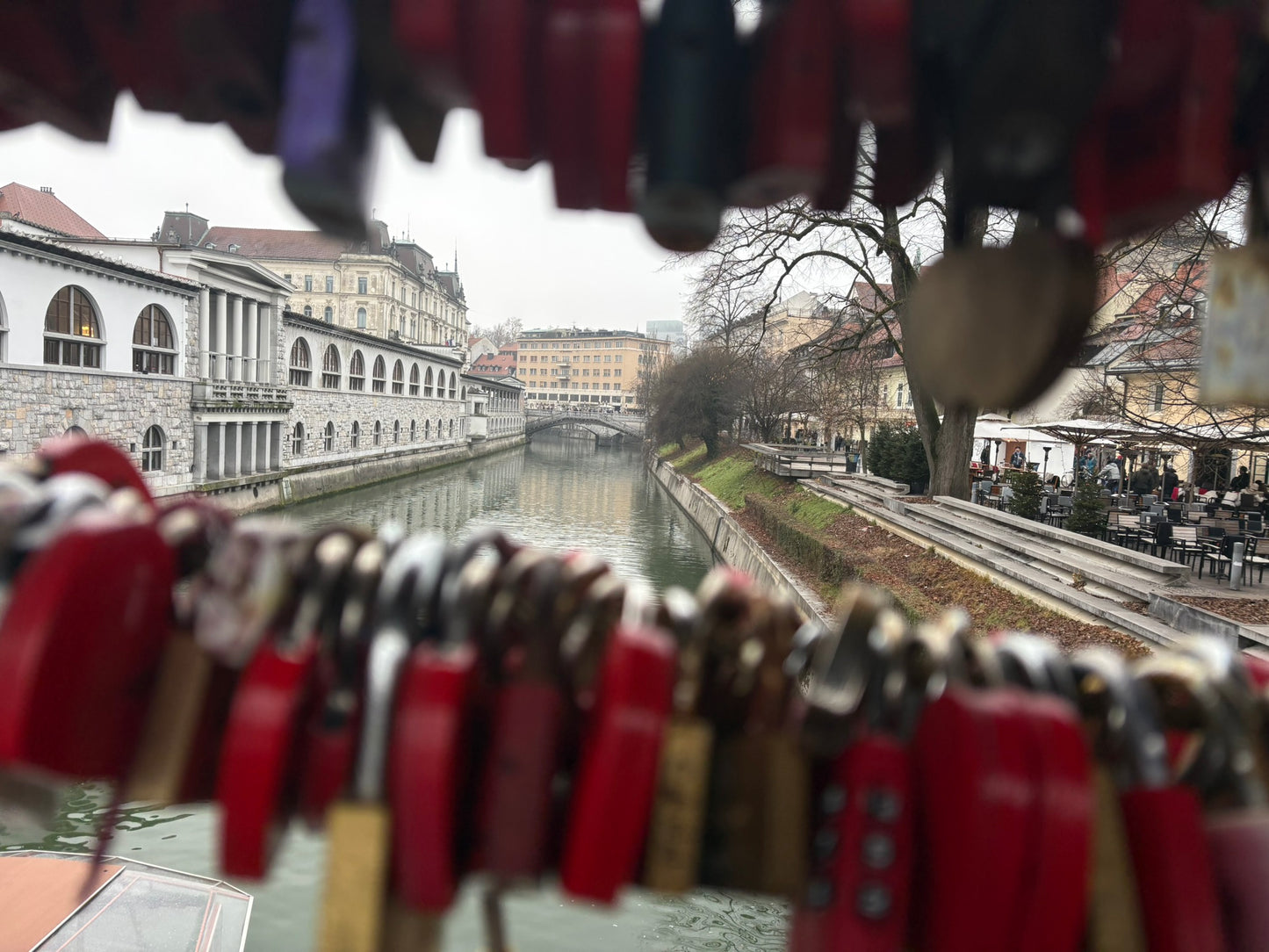 Eine Aufnahme aus der Perspektive eines Betrachters auf einer Brücke, eingerahmt von zahlreichen Liebesschlössern im Vordergrund.

Vordergrund: Eine Vielzahl von bunten, überwiegend roten und messingfarbenen Vorhängeschlössern hängt dicht an dicht an Drahtseilen. Die Schlösser sind leicht unscharf (Bokeh-Effekt), wodurch der Fokus auf den Hintergrund gelenkt wird.

Mittelgrund: Der ruhige Fluss Ljubljanica fließt zentral durch das Bild. Auf der linken Seite erstreckt sich das markante, weiße Arkadengebäude 