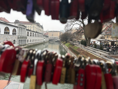 Eine Aufnahme aus der Perspektive eines Betrachters auf einer Brücke, eingerahmt von zahlreichen Liebesschlössern im Vordergrund.

Vordergrund: Eine Vielzahl von bunten, überwiegend roten und messingfarbenen Vorhängeschlössern hängt dicht an dicht an Drahtseilen. Die Schlösser sind leicht unscharf (Bokeh-Effekt), wodurch der Fokus auf den Hintergrund gelenkt wird.

Mittelgrund: Der ruhige Fluss Ljubljanica fließt zentral durch das Bild. Auf der linken Seite erstreckt sich das markante, weiße Arkadengebäude 