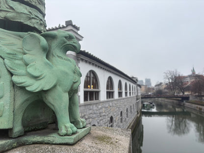 Eine Nahaufnahme einer grün patinierten Drachenskulptur auf einem Steinsockel, die über den Fluss blickt.

Vordergrund: Auf der linken Bildseite steht eine detailliert ausgearbeitete Skulptur eines Drachen aus grüner Bronze. Zu sehen sind die kräftigen Klauen, der geschuppte Körper und ein Teil der majestätisch gefalteten Flügel. Der Drache blickt aufmerksam nach rechts in die Ferne.

Mittelgrund: Direkt hinter dem Drachen erstreckt sich die lange, weiße Arkadenreihe des Zentralmarktes (entworfen von Jože P