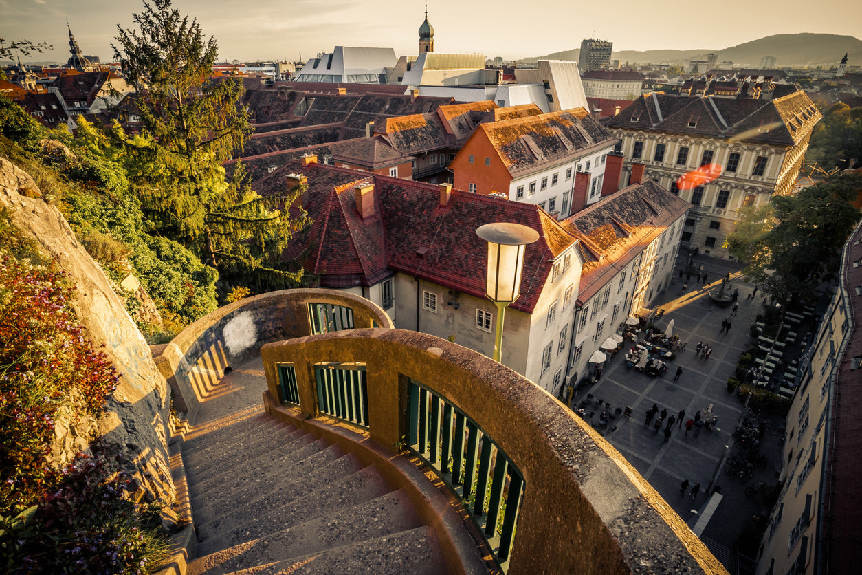 Blick von der Schlossbergtreppe auf den Schloßbergplatz in der Grazer Altstadt bei Sonnenuntergang. Urbanes Stadtpanorama von Graz mit roten Ziegeldächern, historischer Architektur und dem modernen Kunsthaus im Hintergrund – ixpirity: feel the city