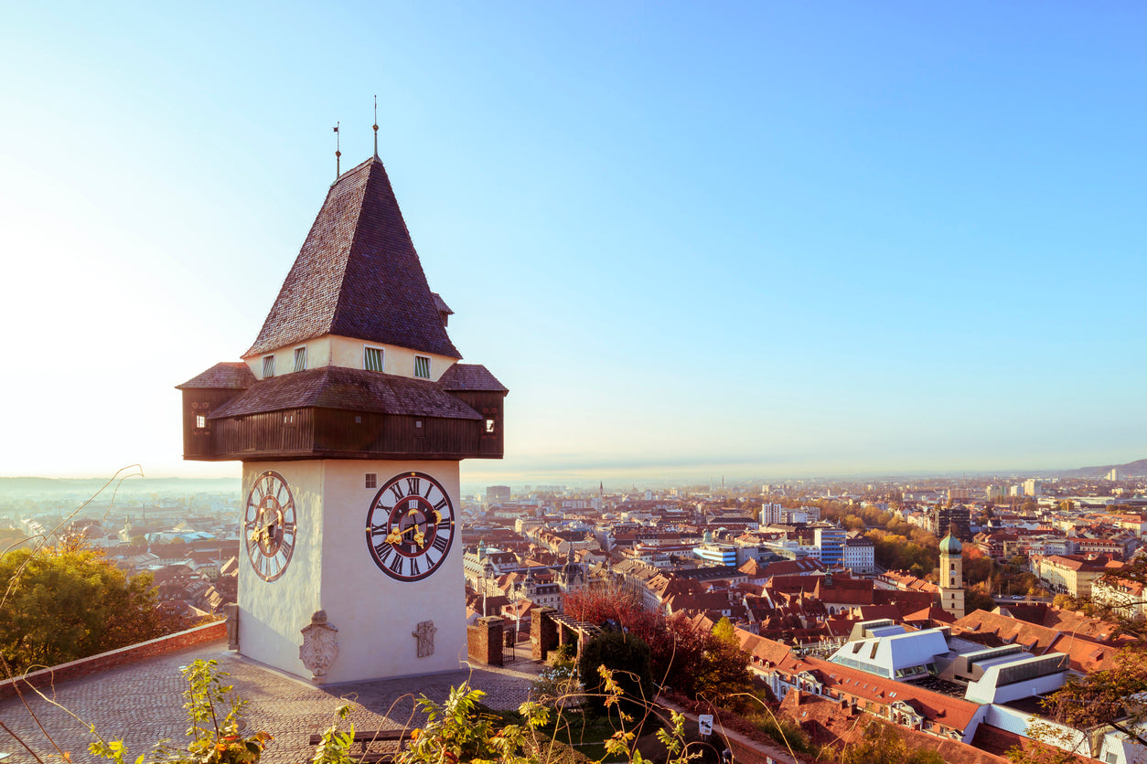 Der berühmte Grazer Uhrturm auf dem Schlossberg bei strahlendem Sonnenschein. Das Bild zeigt das historische Wahrzeichen der Steiermark mit Blick über das weitläufige Stadtpanorama von Graz und die umliegende Hügellandschaft.