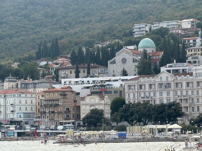 Blick über Opatija von der Badebucht Slatina aus: Grandhotels und alte Villen am Hang, darüber die grüne Kuppel der Kirche Mariä Verkündigung und die bewaldeten Hänge der Učka.
