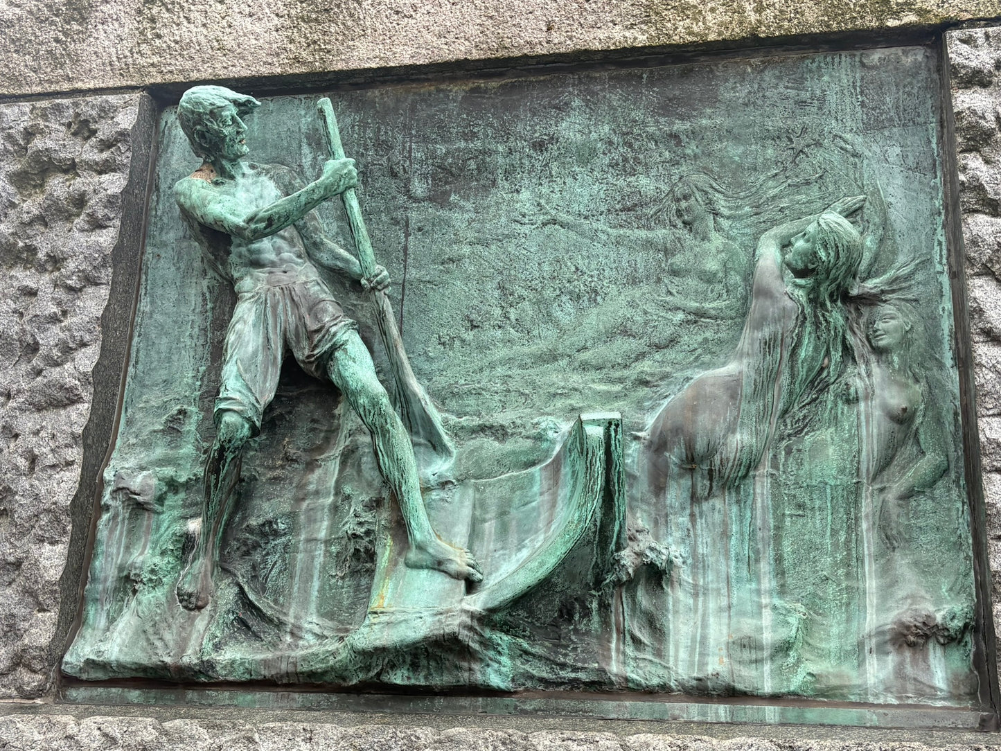 Ein detailreiches, grün patiniertes Bronze-Relief, eingelassen in rauen Stein. Es zeigt links einen Mann, der mit einer Stange auf einem Boot steht, während rechts mehrere weibliche Figuren (Wassernymphen) aus den Wellen nach ihm greifen.