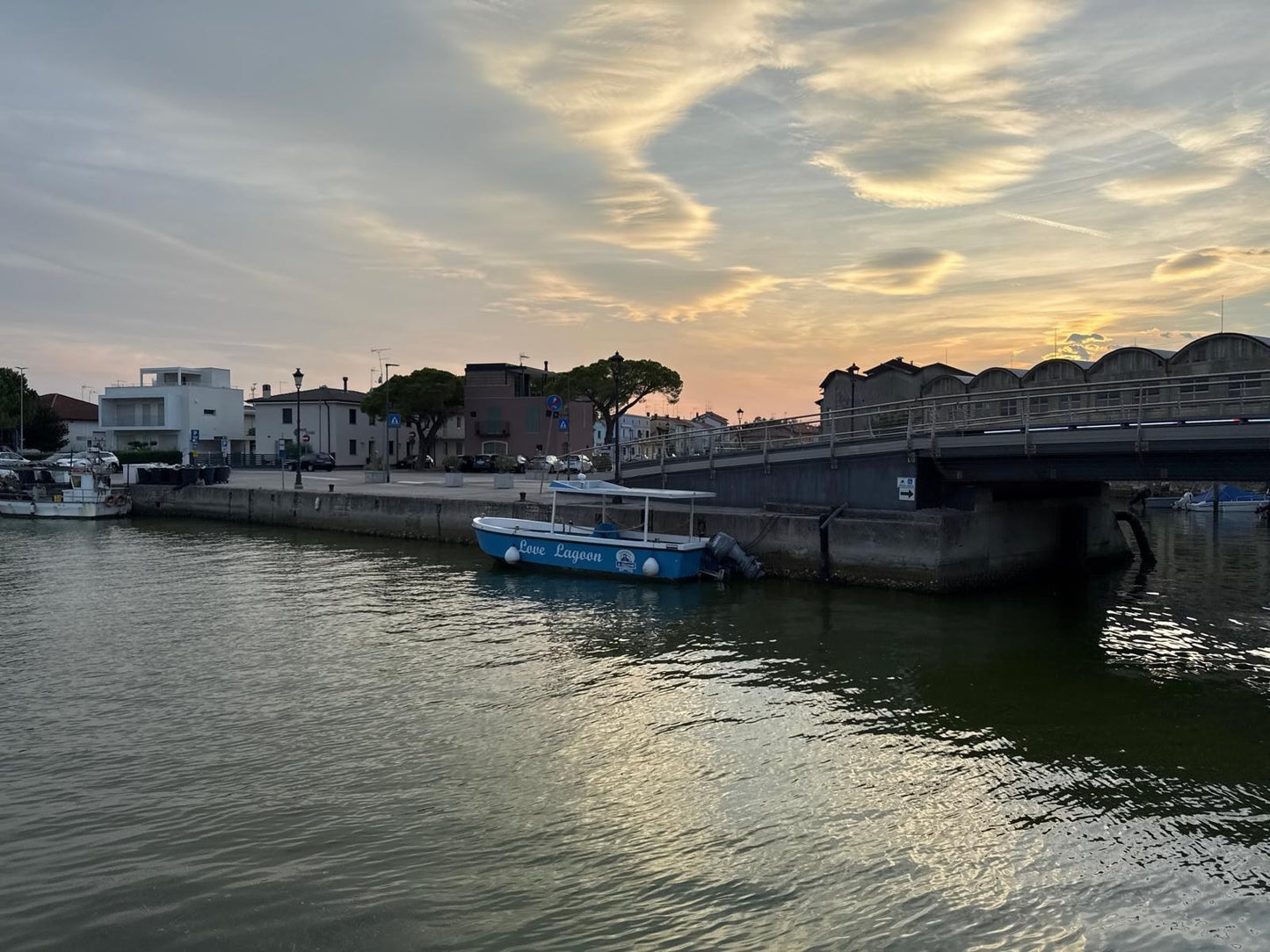 Der Kanal von Marano Lagunare bei Sonnenuntergang mit einem blauen Ausflugsboot an der Brücke und goldenem Licht auf dem Wasser