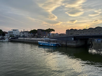 Der Kanal von Marano Lagunare bei Sonnenuntergang mit einem blauen Ausflugsboot an der Brücke und goldenem Licht auf dem Wasser