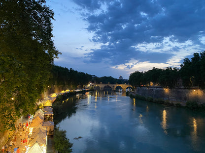 Il Tevere al crepuscolo con le bancarelle sulla riva e la cupola di San Pietro