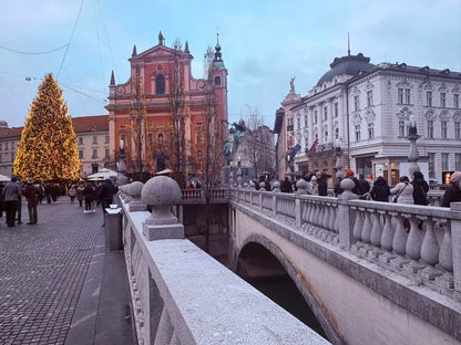 Stimmungsvolle Aufnahme des Prešeren-Platzes in Ljubljana zur Weihnachtszeit, mit einem großen beleuchteten Weihnachtsbaum, der rosa Franziskanerkirche und Menschen auf den Drei Brücken.